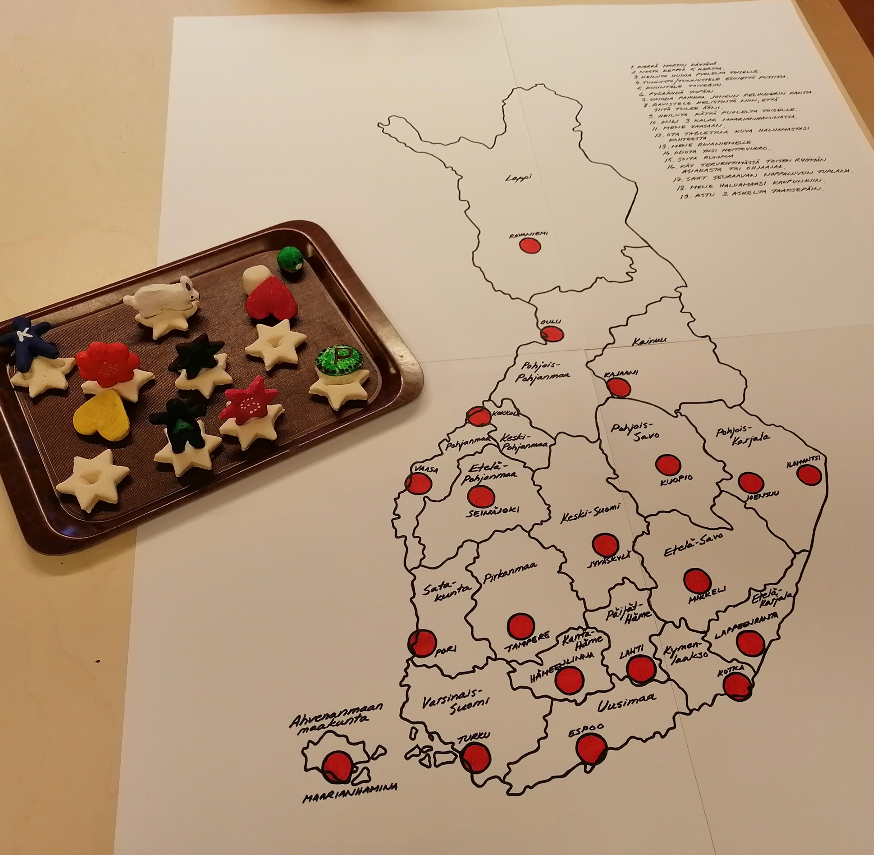 Paperille piirretty Suomen kartta, jonka päällä punaisia merkkejä maakuntien päällä. Karttapiirroksen vieressä musta tarjotin, jolla erivärisiä pelimerkkejä.
